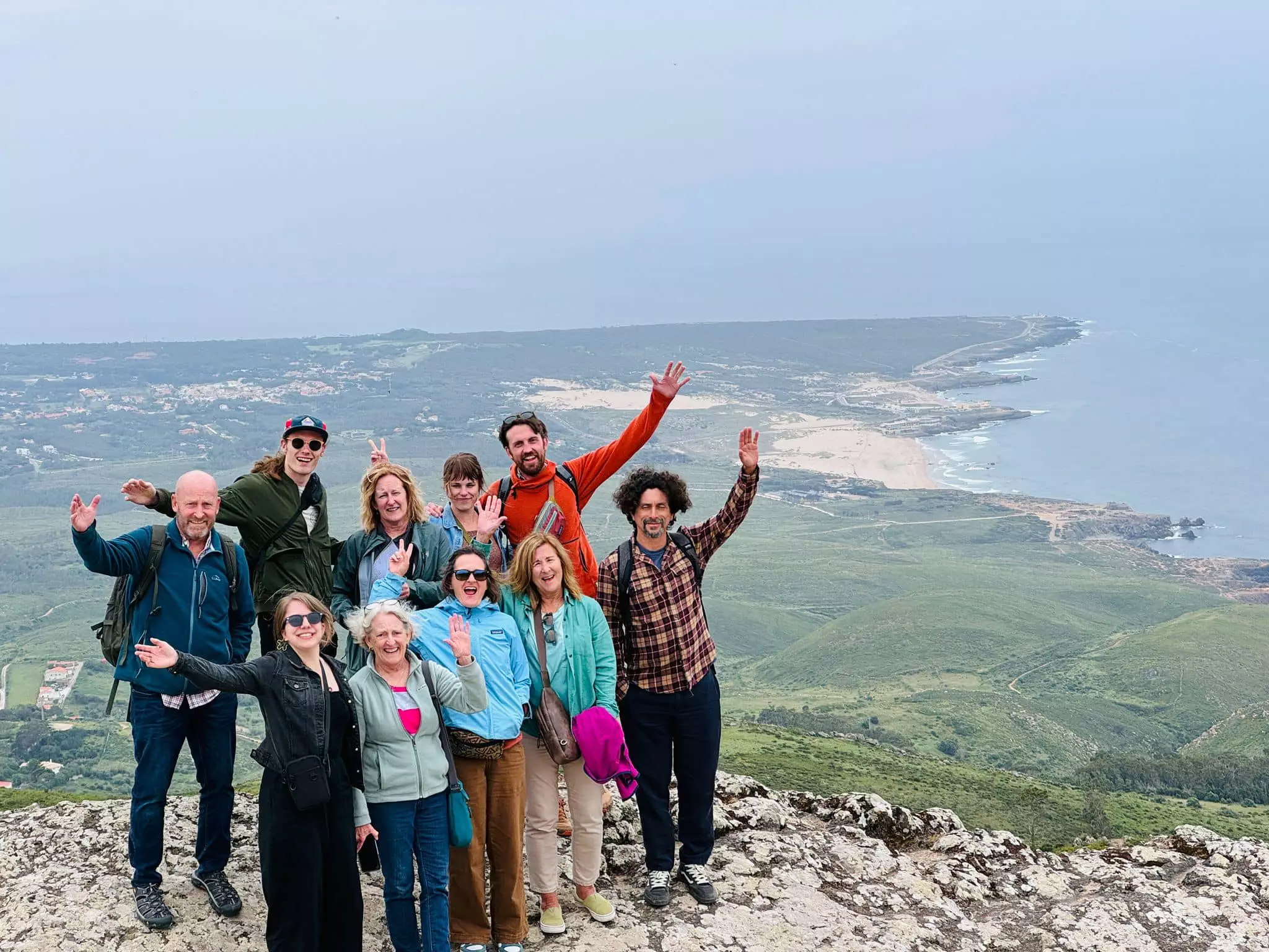 A joyful group of hikers poses at the top of a cliff with panoramic views of the coast near Sintra, Portugal. Part of a Yes, You Deserve tour, they celebrate the moment with raised hands and big smiles, surrounded by stunning green hills and ocean scenery.