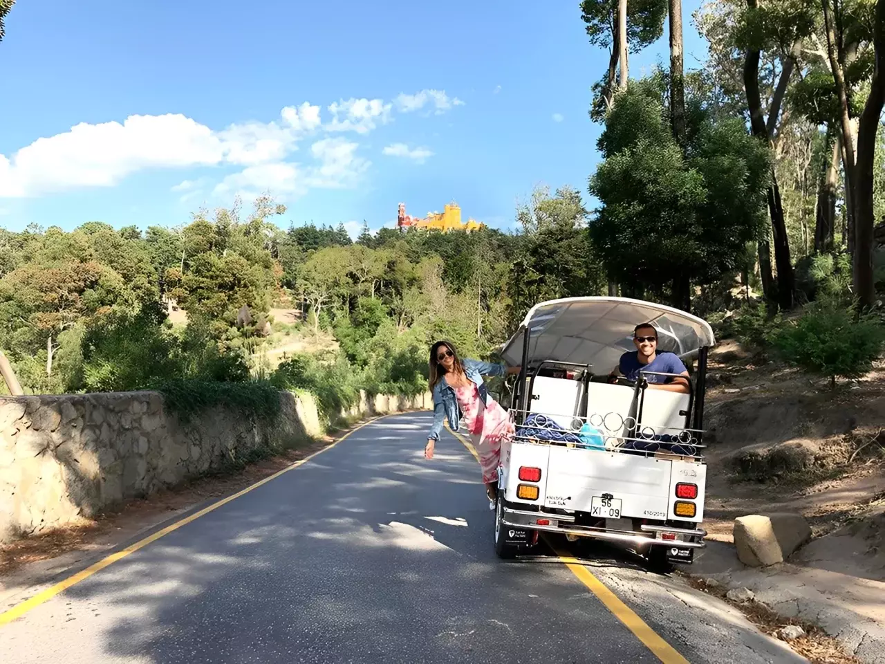 A white tuk-tuk from Yes, You Deserve drives through a scenic forest road in Sintra, with smiling tourists enjoying the ride. In the distance, the colorful Pena Palace peeks through the trees under a bright blue sky.