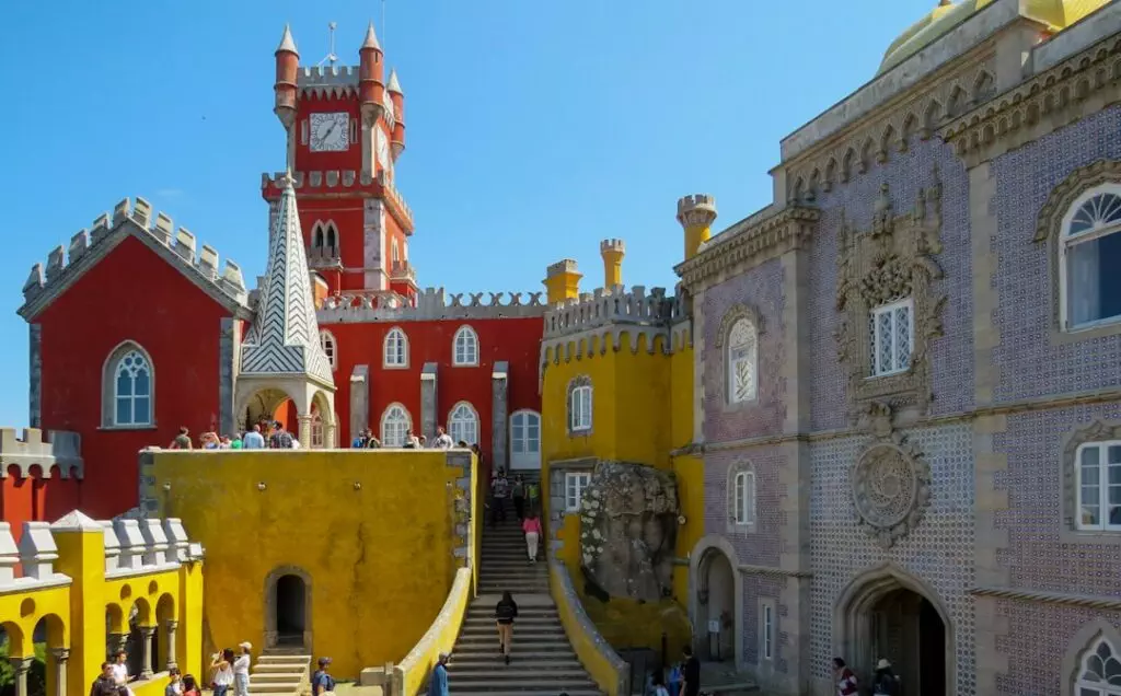 Panoramic view of the vibrant and eclectic architecture of Pena Palace in Sintra, Portugal, with tourists exploring its colorful red, yellow, and tiled façades — a highlight of Yes, You Deserve’s private tuk tuk tours.