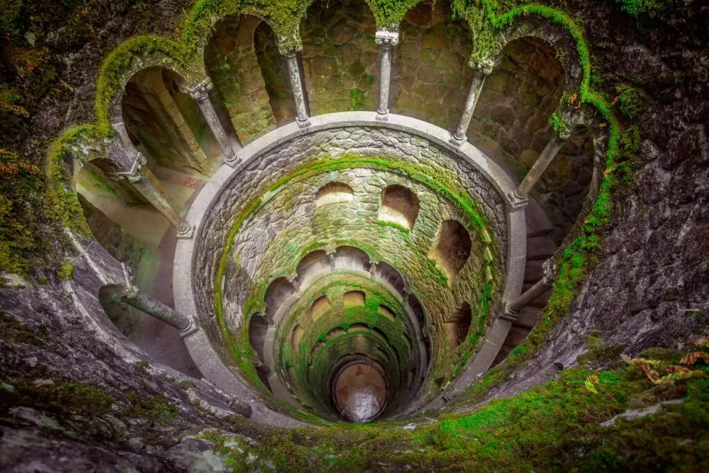 Initiation Well at Quinta da Regaleira in Sintra, Portugal – a mystical underground spiral staircase used for Masonic and Rosicrucian symbolic rituals, surrounded by moss-covered stone.