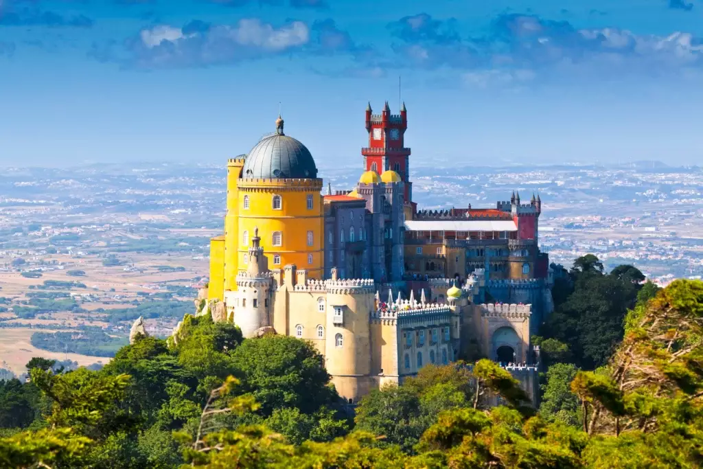 Panoramic view of the colorful Pena Palace in Sintra, Portugal, perched on a hilltop with stunning landscape in the background under a bright blue sky.