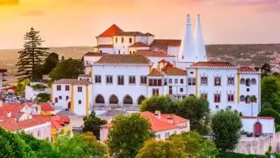 Sunset view of the National Palace of Sintra with its iconic twin chimneys, surrounded by colorful rooftops and lush greenery in the historic center of Sintra, Portugal.
