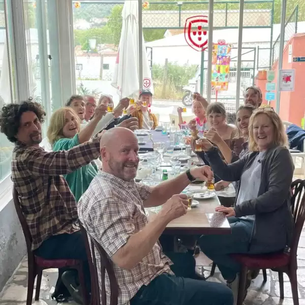 A joyful group of tourists from a Yes, You Deserve tour raises their glasses for a toast during a traditional Portuguese lunch in Sintra. Seated around a long table, they celebrate the experience with smiles and good company inside a cozy local restaurant.