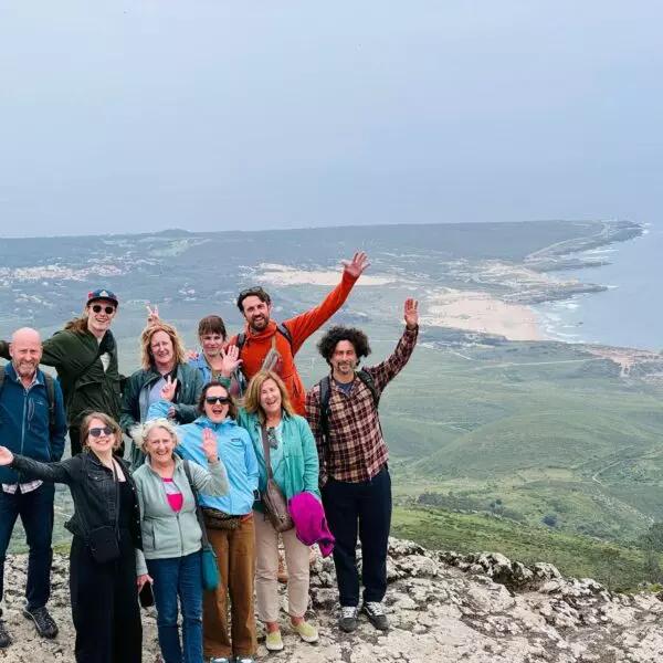 A joyful group of hikers poses at the top of a cliff with panoramic views of the coast near Sintra, Portugal. Part of a Yes, You Deserve tour, they celebrate the moment with raised hands and big smiles, surrounded by stunning green hills and ocean scenery.