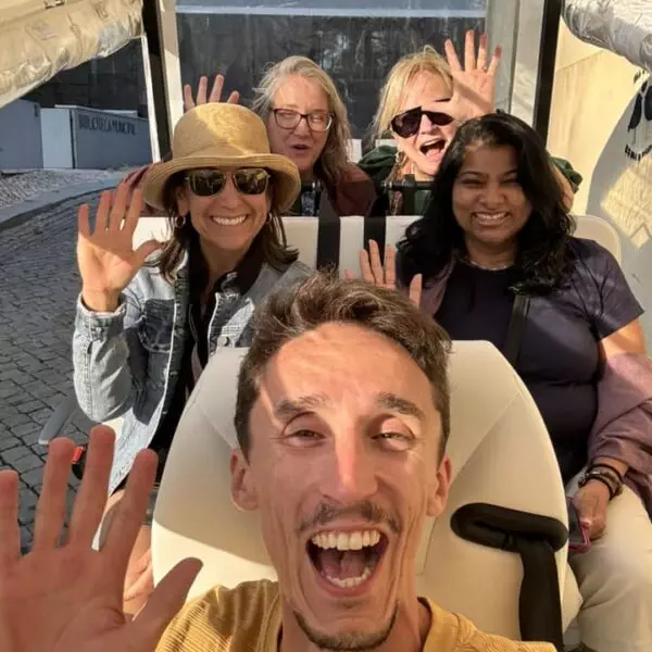 Tour guide Daniel Ponce smiles widely with a group of five excited women inside a white tuk-tuk during a Yes, You Deserve tour in Sintra. Everyone is waving and enjoying the golden afternoon sunlight, capturing the joy of the moment.