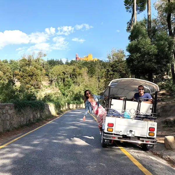 A white tuk-tuk from Yes, You Deserve drives through a scenic forest road in Sintra, with smiling tourists enjoying the ride. In the distance, the colorful Pena Palace peeks through the trees under a bright blue sky.