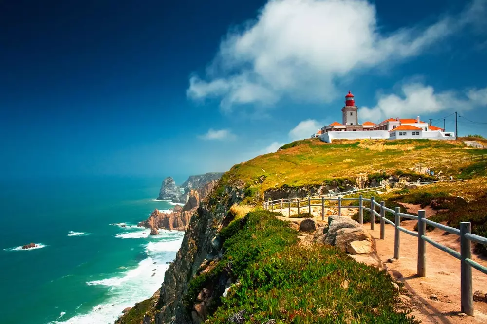 Scenic view of Cabo da Roca, the westernmost point of continental Europe, featuring dramatic cliffs, the Atlantic Ocean, and a historic lighthouse under a vibrant blue sky. A popular stop on the Yes, You Deserve Sintra and coastal tour.