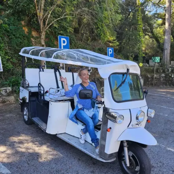 A smiling woman poses cheerfully in the driver’s seat of a white electric tuk-tuk parked in a scenic forested area in Sintra, Portugal. Captured during a Yes, You Deserve tour, the moment radiates fun and adventure.