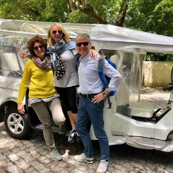 Three happy tourists pose in front of a white electric tuk-tuk during a Yes, You Deserve tour in Sintra, Portugal. The group is smiling under the trees, ready to explore the city in a fun and eco-friendly way.